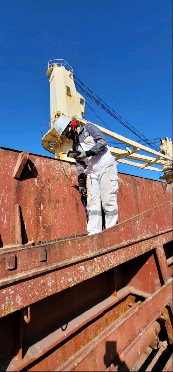 A Moim surveyor carrying out ultrasonic testing of a bulk carrier's hatch covers, standing on the hatch coaming with a deck crane behind.